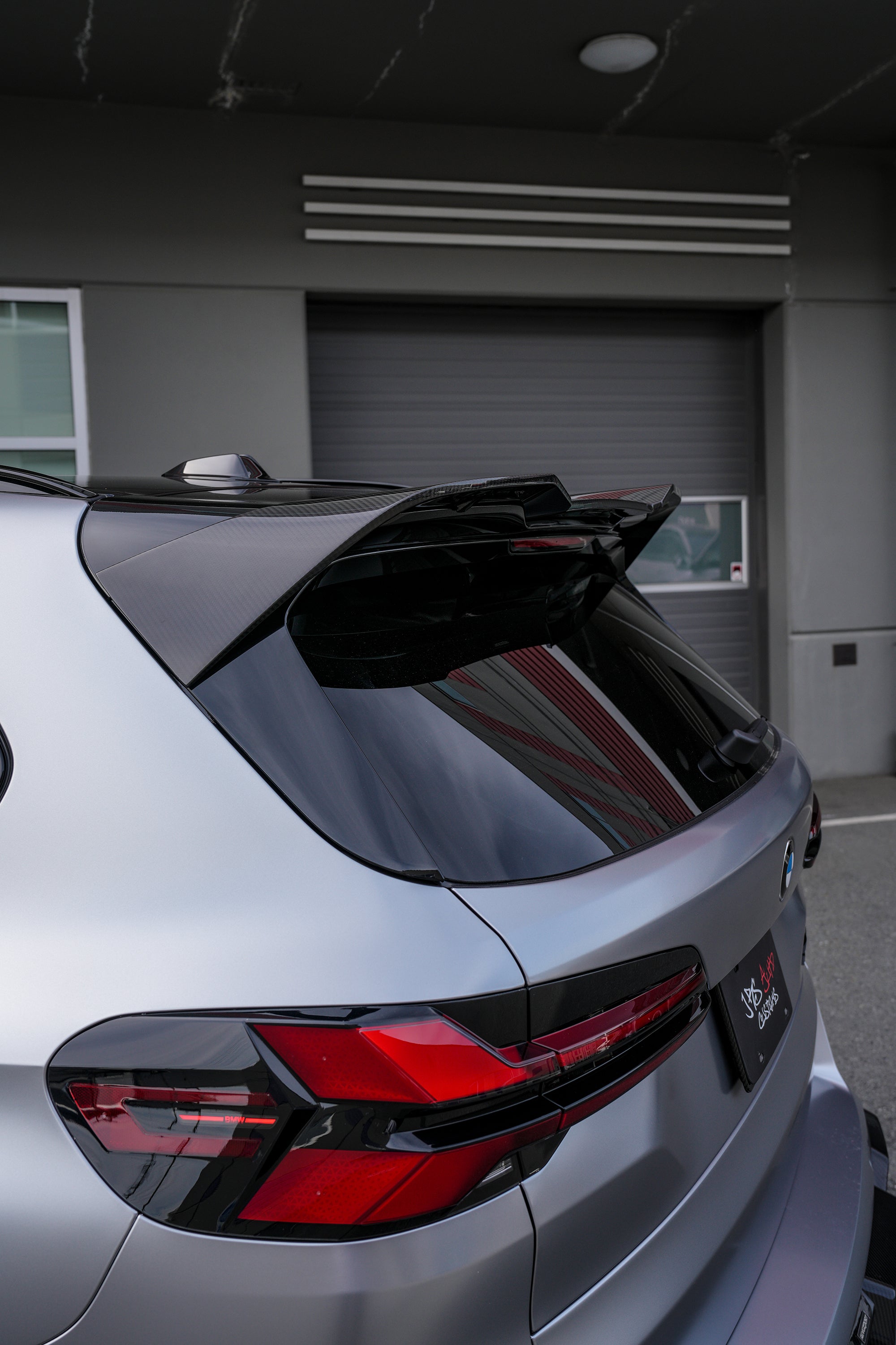 Gray BMW X5M featuring a REVOZPORT carbon fiber body kit with aggressive front lip, side skirts, and sporty aero styling parked outside a workshop.