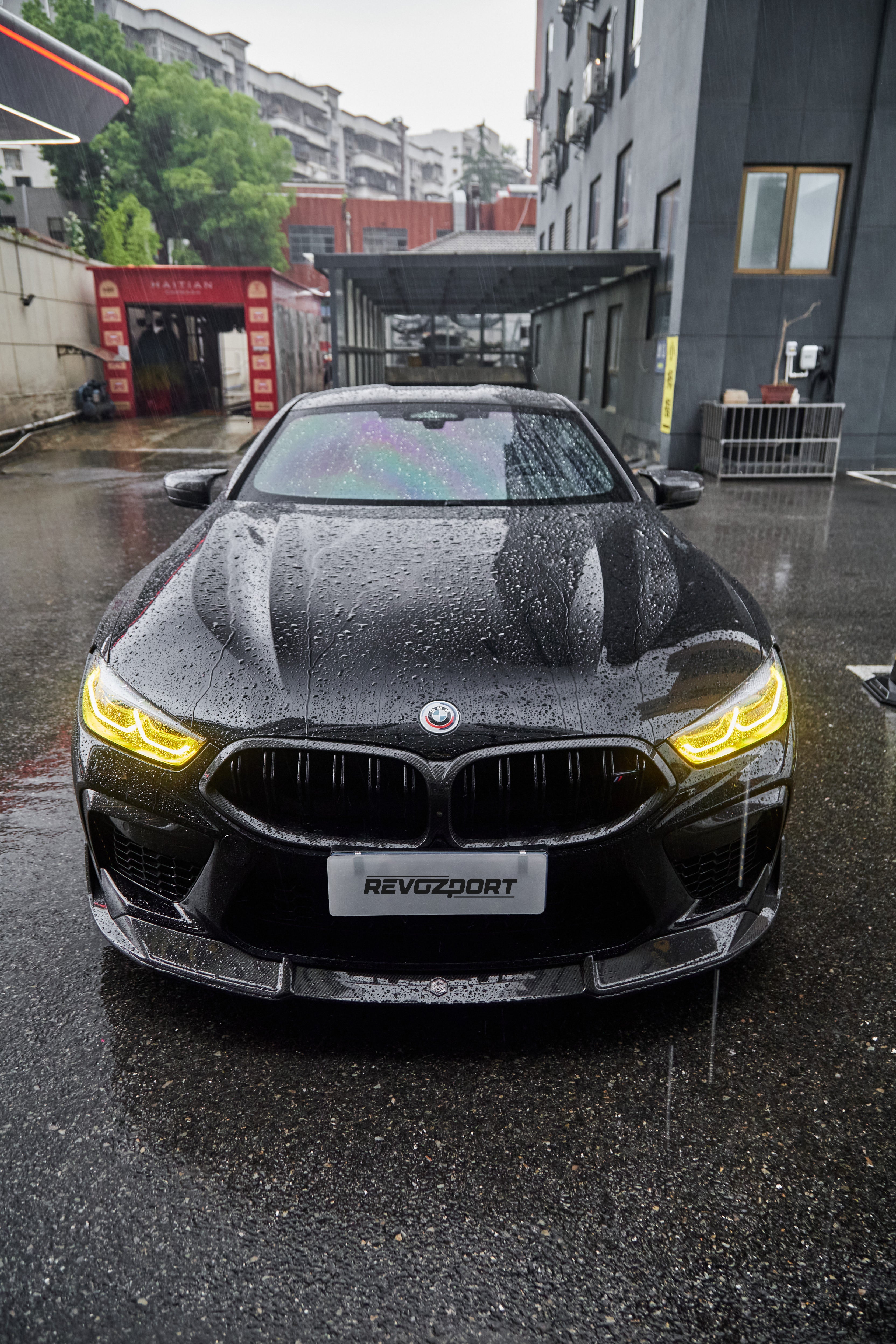 Rain-covered BMW front view with yellow headlights, carbon fiber hood, front lip, and aggressive aero styling on wet pavement.