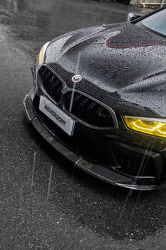 Rain-covered BMW front close-up with yellow headlights, carbon fiber hood, front lip, and aggressive aero styling.