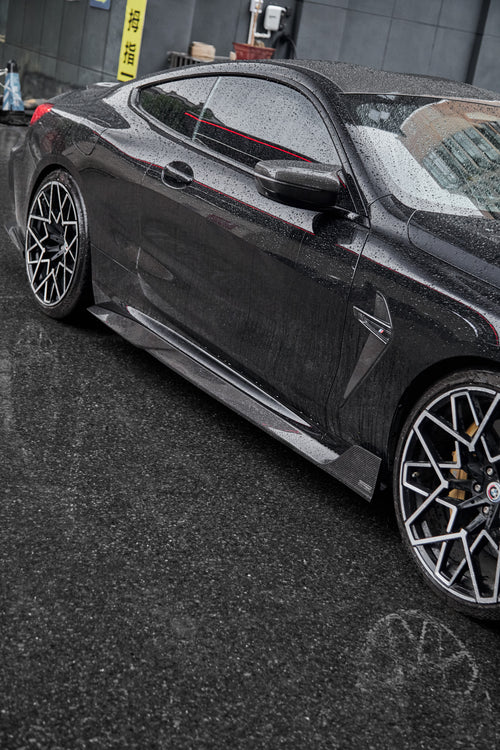 Rain-covered BMW side profile with carbon fiber side skirts, aggressive wheel fitment, and sleek performance styling on wet pavement.