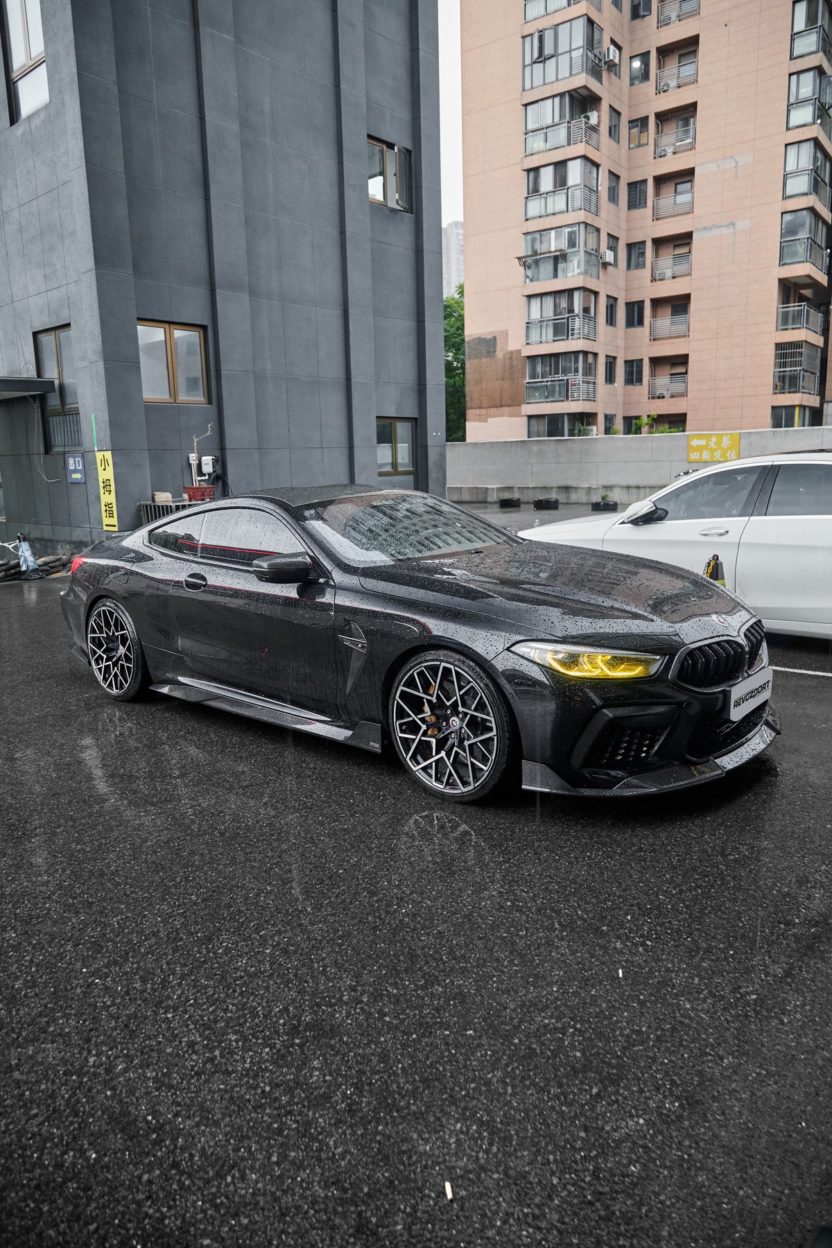 Rain-covered BMW with carbon fiber body kit, yellow headlights, and aggressive stance parked on wet pavement in an urban setting.