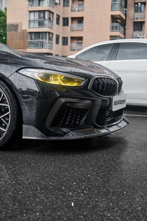 Rain-covered BMW front end with carbon fiber lip, yellow headlights, and aggressive aero styling on wet pavement.