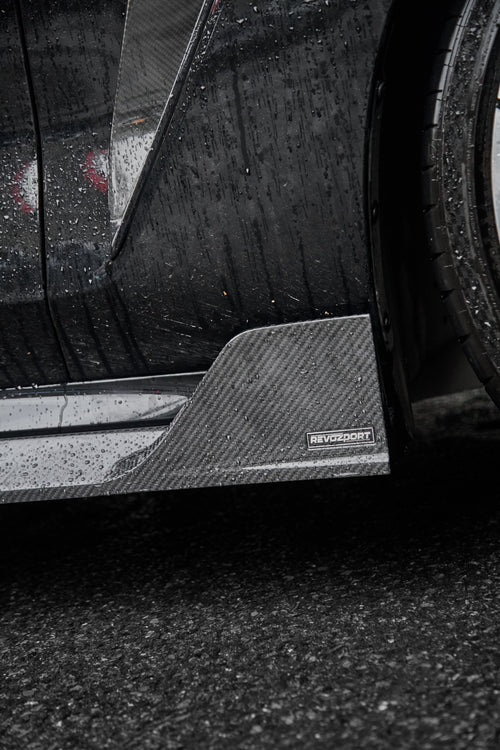 Close-up of a rain-covered carbon fiber side skirt and aero fin, highlighting glossy weave texture and precision detailing.