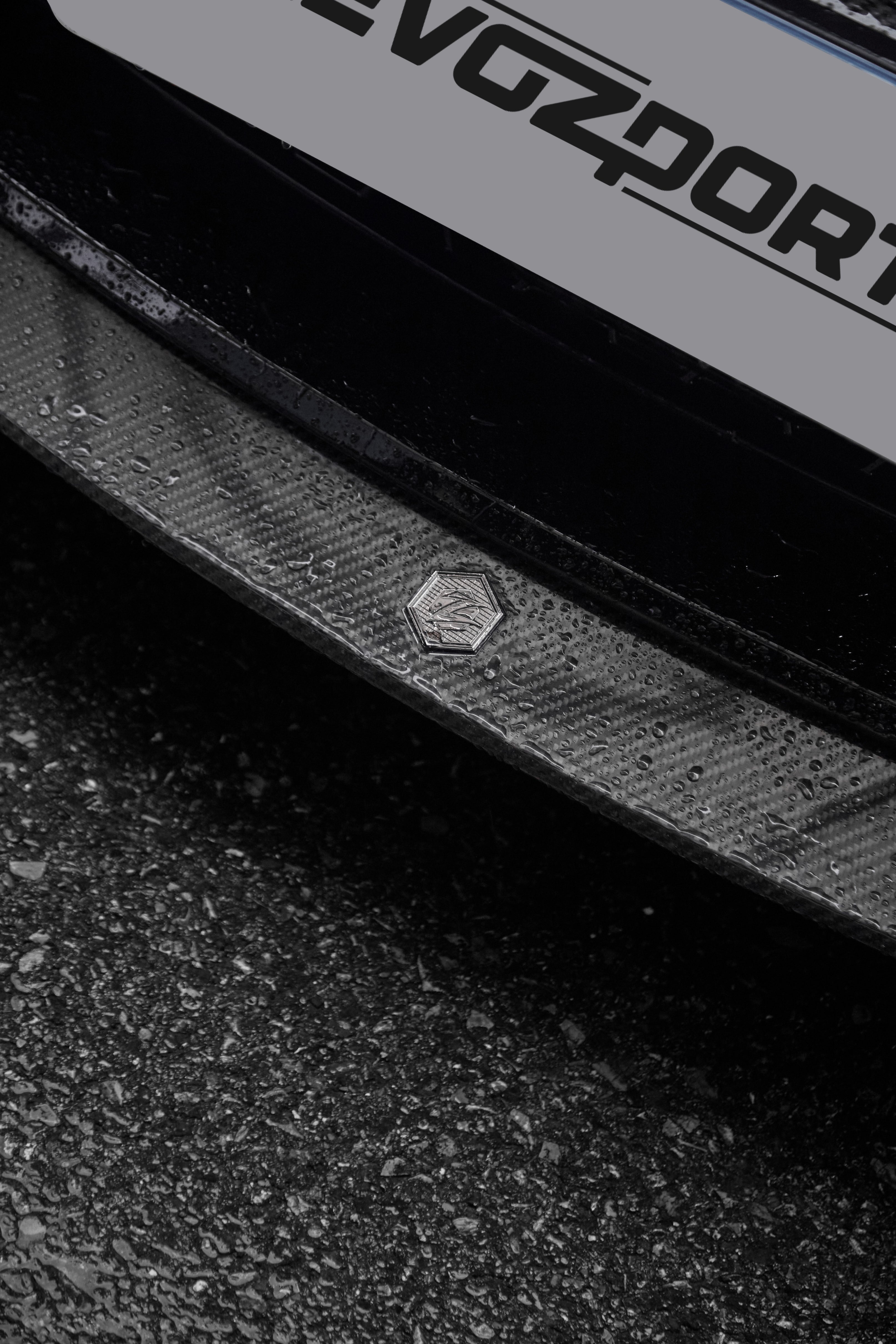 Close-up of a rain-covered carbon fiber front lip, highlighting glossy weave texture, precision badge detail, and wet pavement.