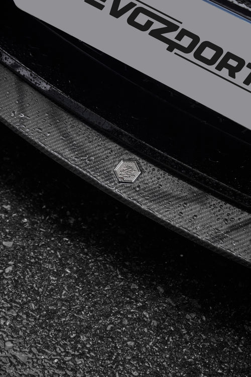 Close-up of a rain-covered carbon fiber front lip, highlighting glossy weave texture, precision badge detail, and wet pavement.