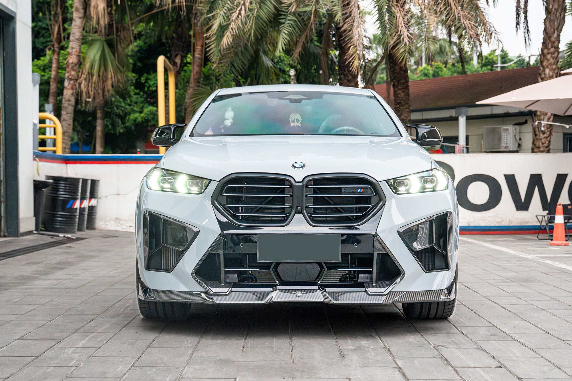 Front-facing view of a white BMW featuring aggressive carbon fiber front aero, illuminated headlights, sharp intake details, and performance-focused styling.
