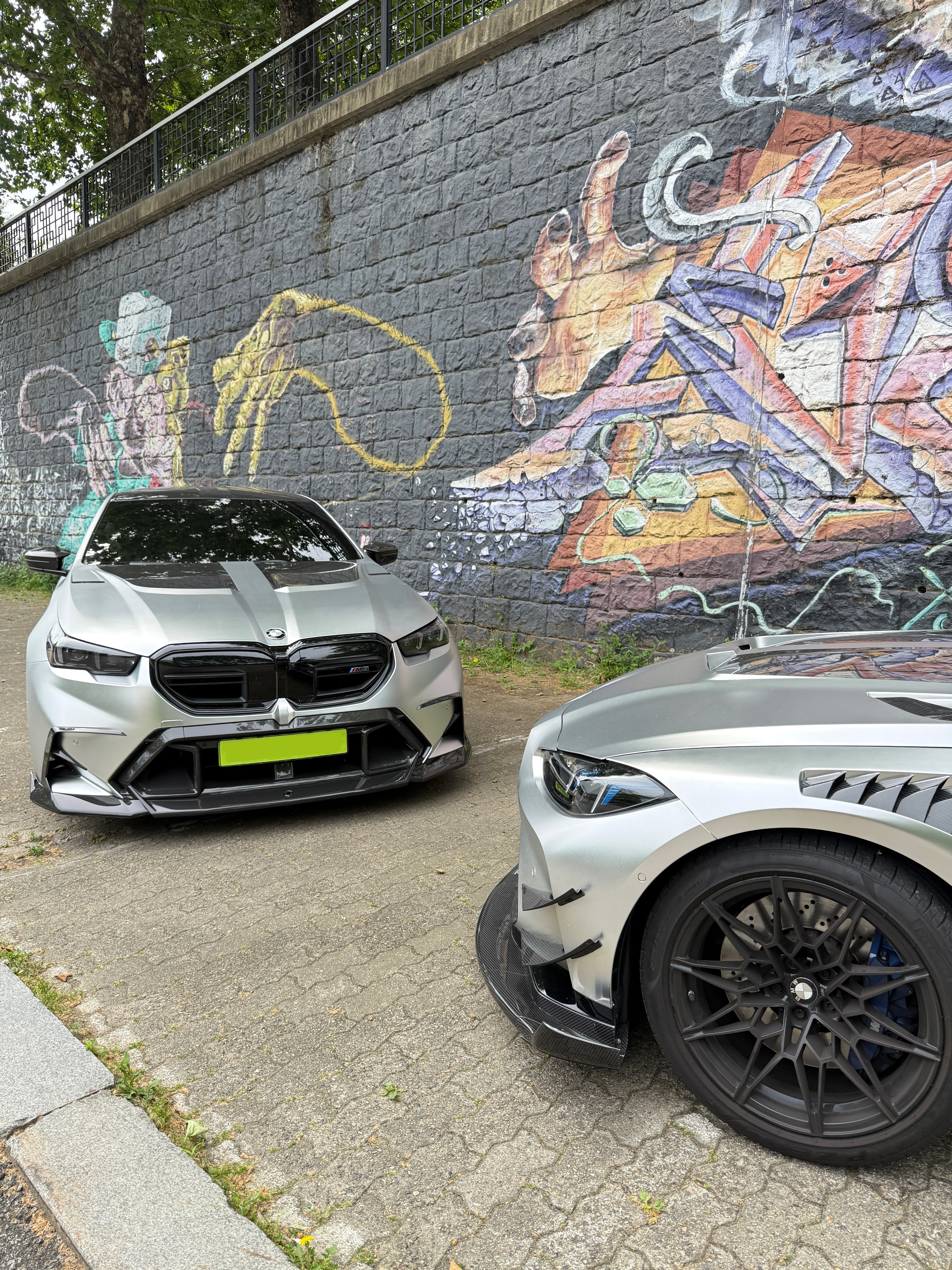 Two BMWs with carbon fiber aero kits parked beside a colorful graffiti wall, highlighting aggressive front styling and urban atmosphere.
