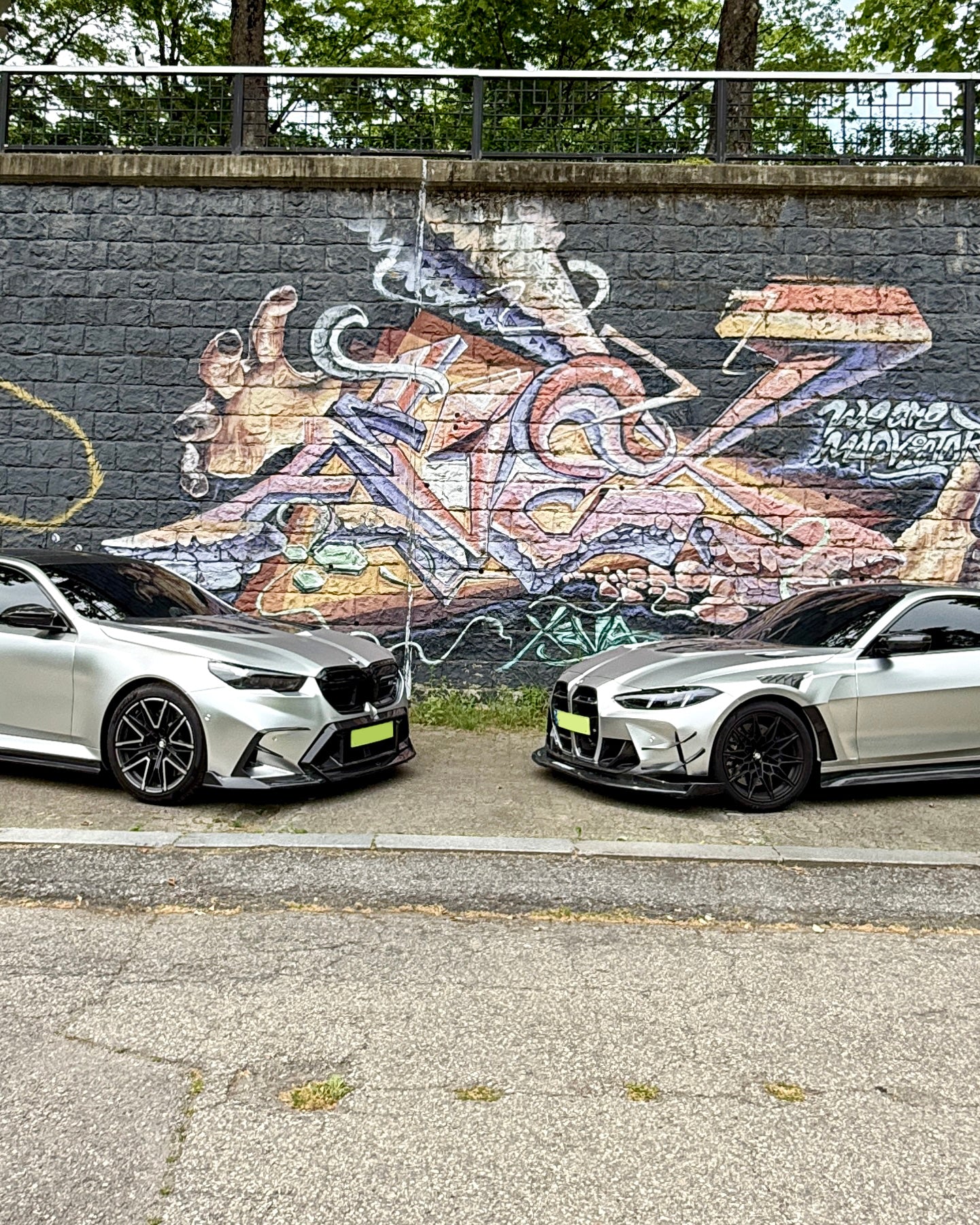 Two BMWs with carbon fiber aero kits parked face-to-face beside a colorful graffiti wall in an urban street setting.