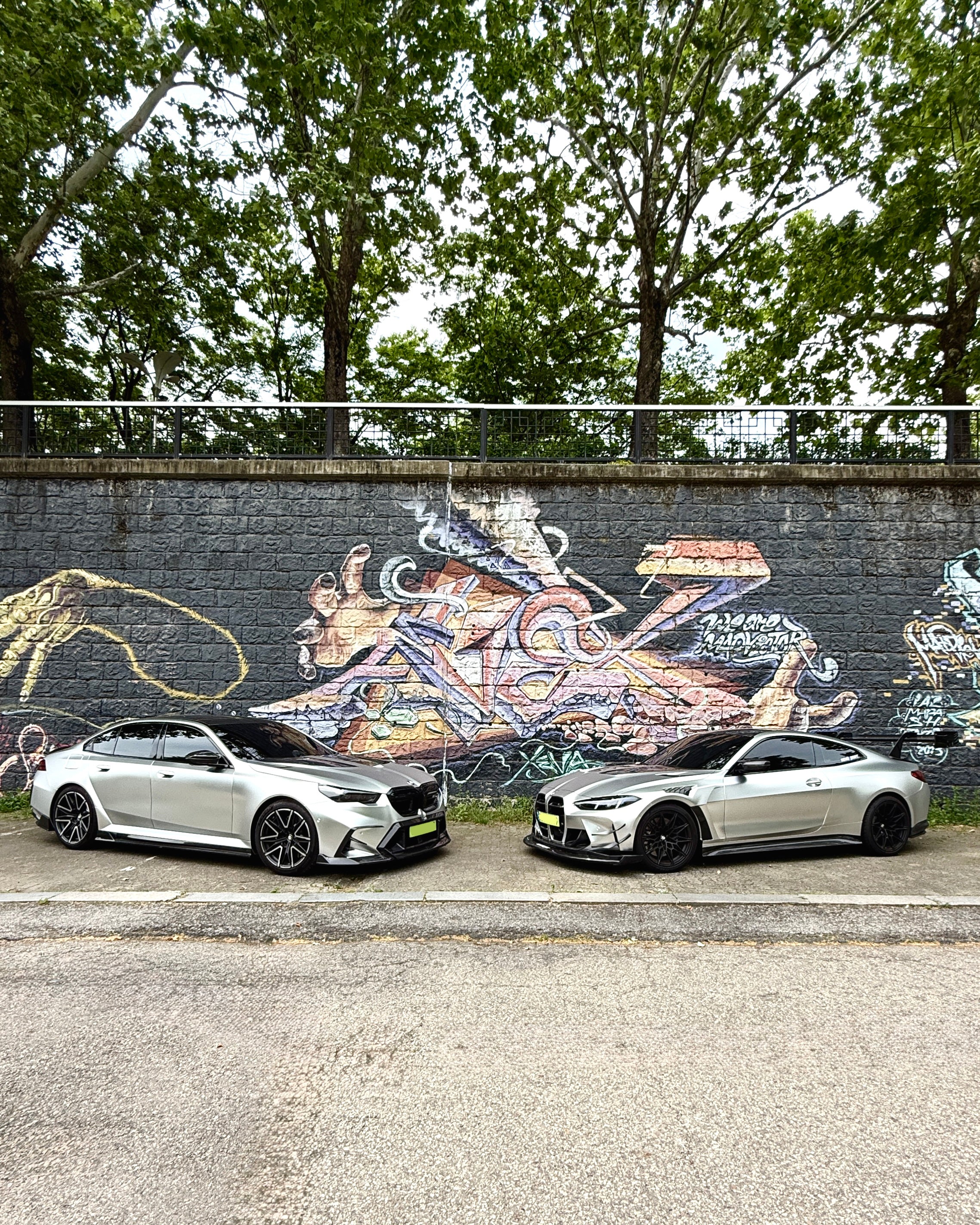Two BMWs with carbon fiber aero kits parked side by side against a colorful graffiti wall in an urban street setting.
