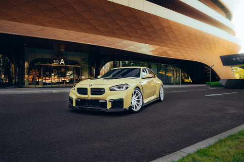 Yellow BMW with carbon fiber body kit parked outside a modern building, highlighting front lip, side skirts, and white wheels.