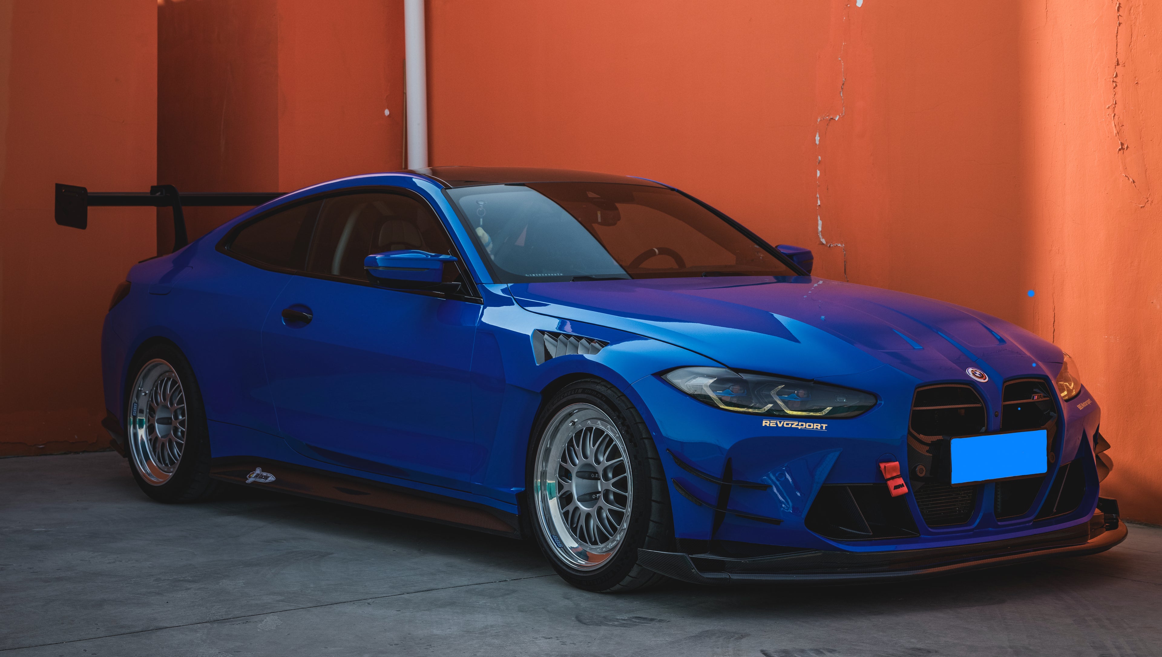 Blue BMW G82 M4 with carbon fiber front splitter, side skirts, rear wing, and aftermarket wheels parked in a modern industrial courtyard with colorful architectural walls.