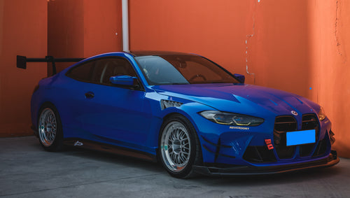 Blue BMW G82 M4 with carbon fiber front splitter, side skirts, rear wing, and aftermarket wheels parked in a modern industrial courtyard with colorful architectural walls.