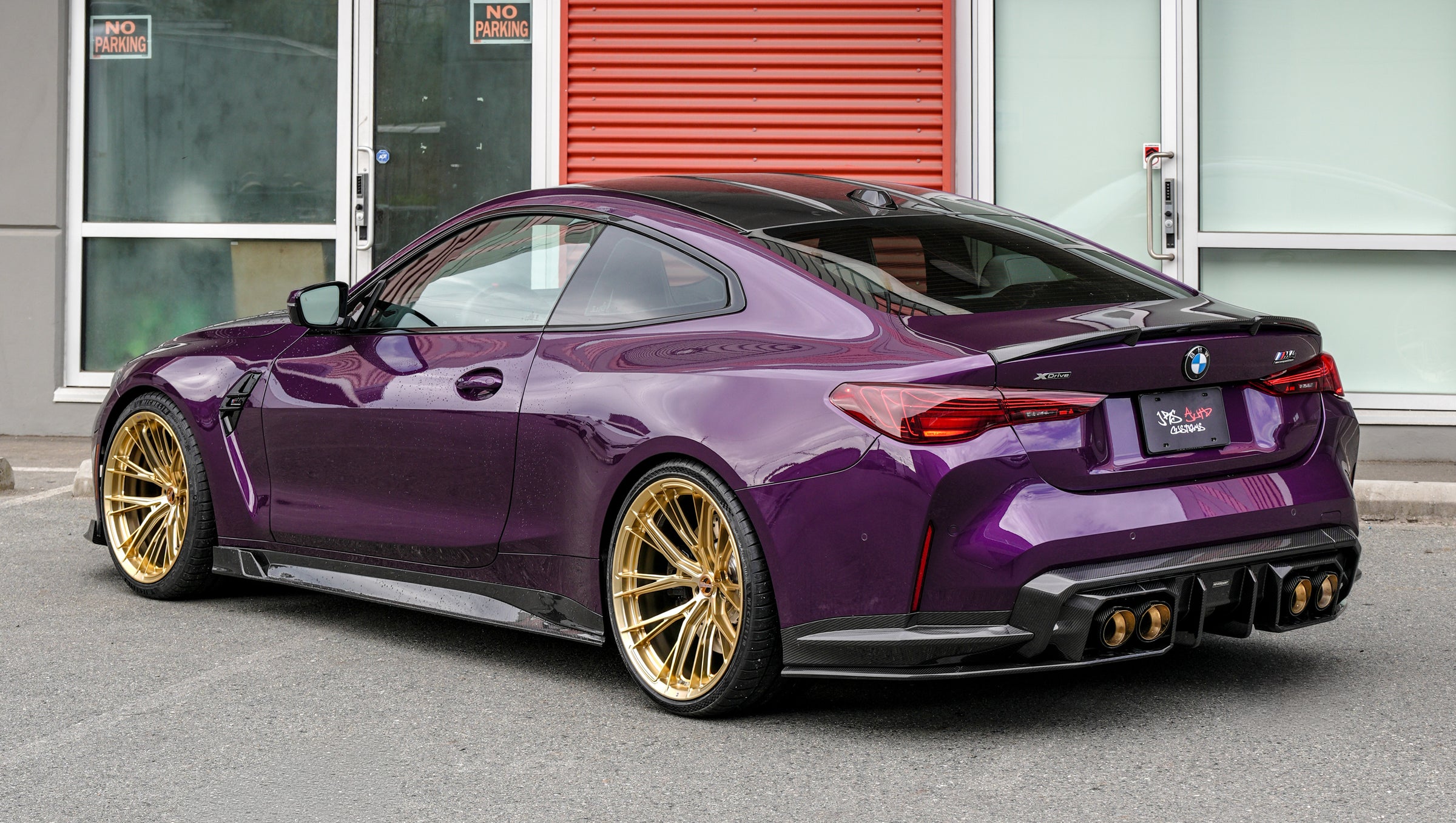 Purple BMW G82 M4 featuring a REVOZPORT carbon fiber body kit with front lip, side skirts, vented fenders, and gold wheels parked outside a modern industrial building.