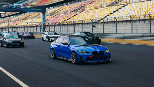 Blue BMW G80 M3 with REVOZPORT carbon fiber front splitter, side skirts, vented fenders, and large rear wing parked in a landscaped outdoor plaza.