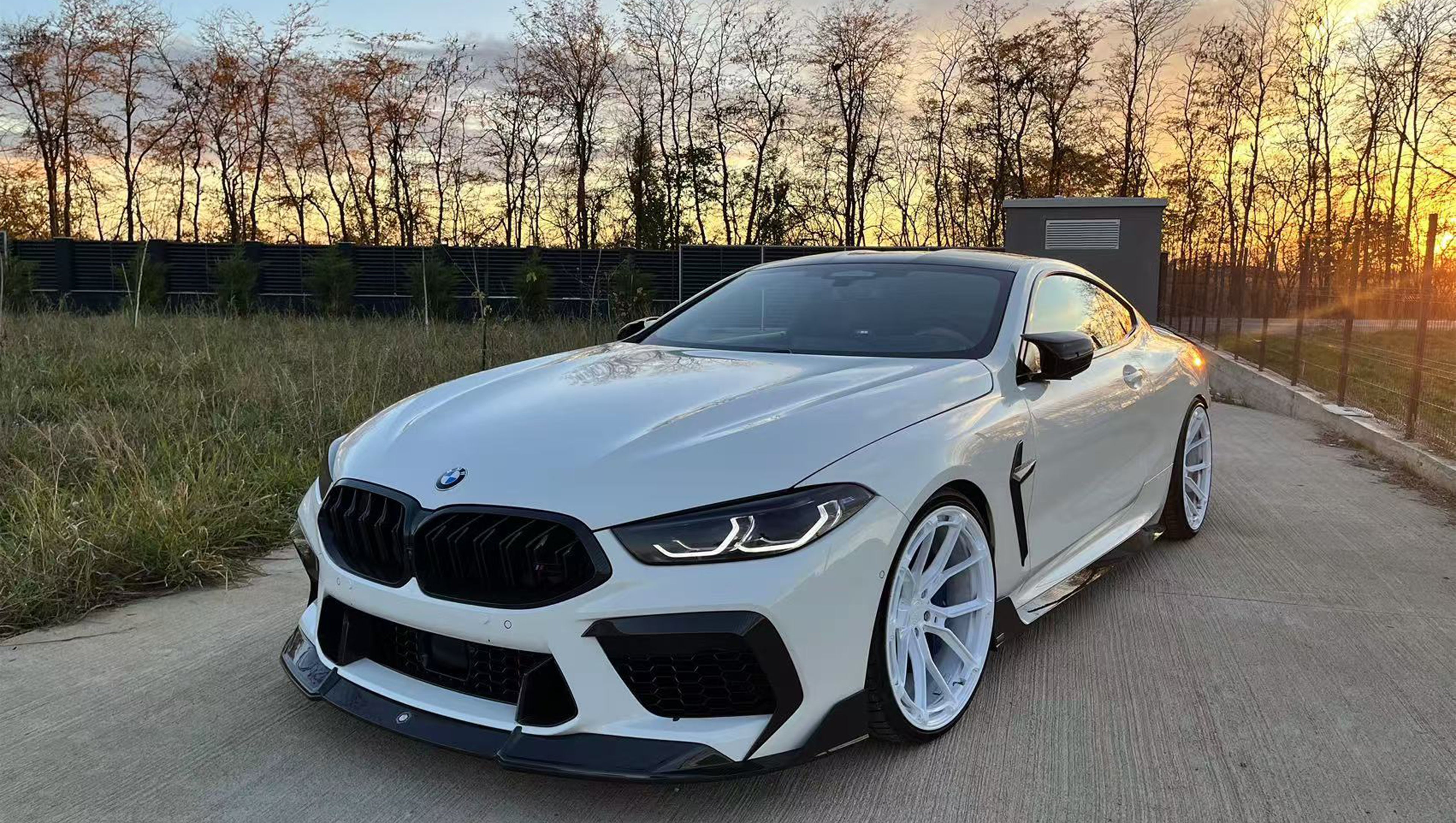 White BMW F92 M8 featuring a REVOZPORT carbon fiber front lip, side skirts, black front grille, and white multi-spoke forged wheels parked at sunset on an open road.