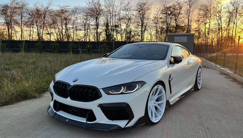 White BMW F92 M8 featuring a REVOZPORT carbon fiber front lip, side skirts, black front grille, and white multi-spoke forged wheels parked at sunset on an open road.