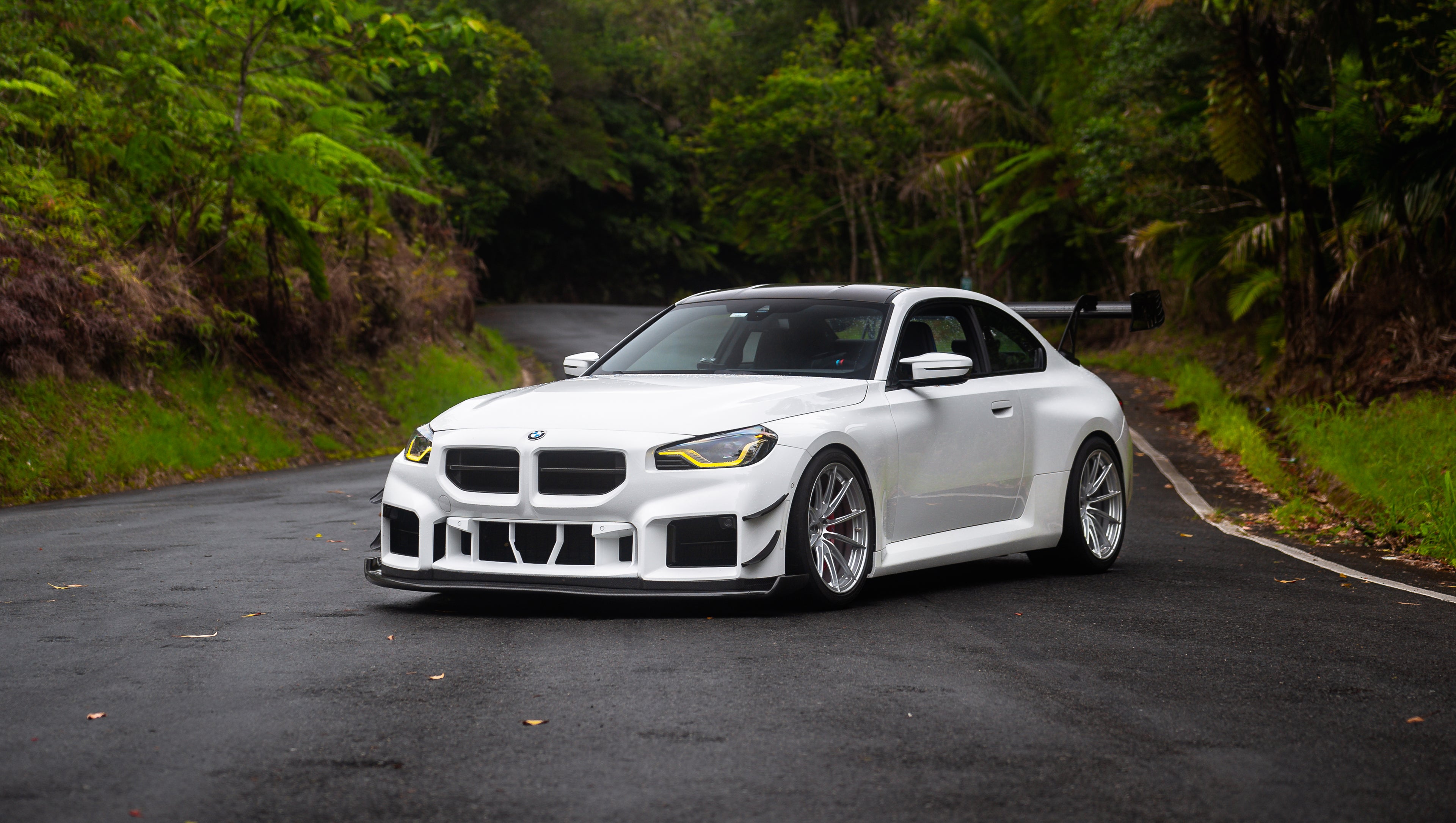 White widebody BMW G82 M4 with aggressive front bumper, canards, deep front splitter, large rear wing, and yellow LED headlights parked on a wet road surrounded by dense tropical forest.