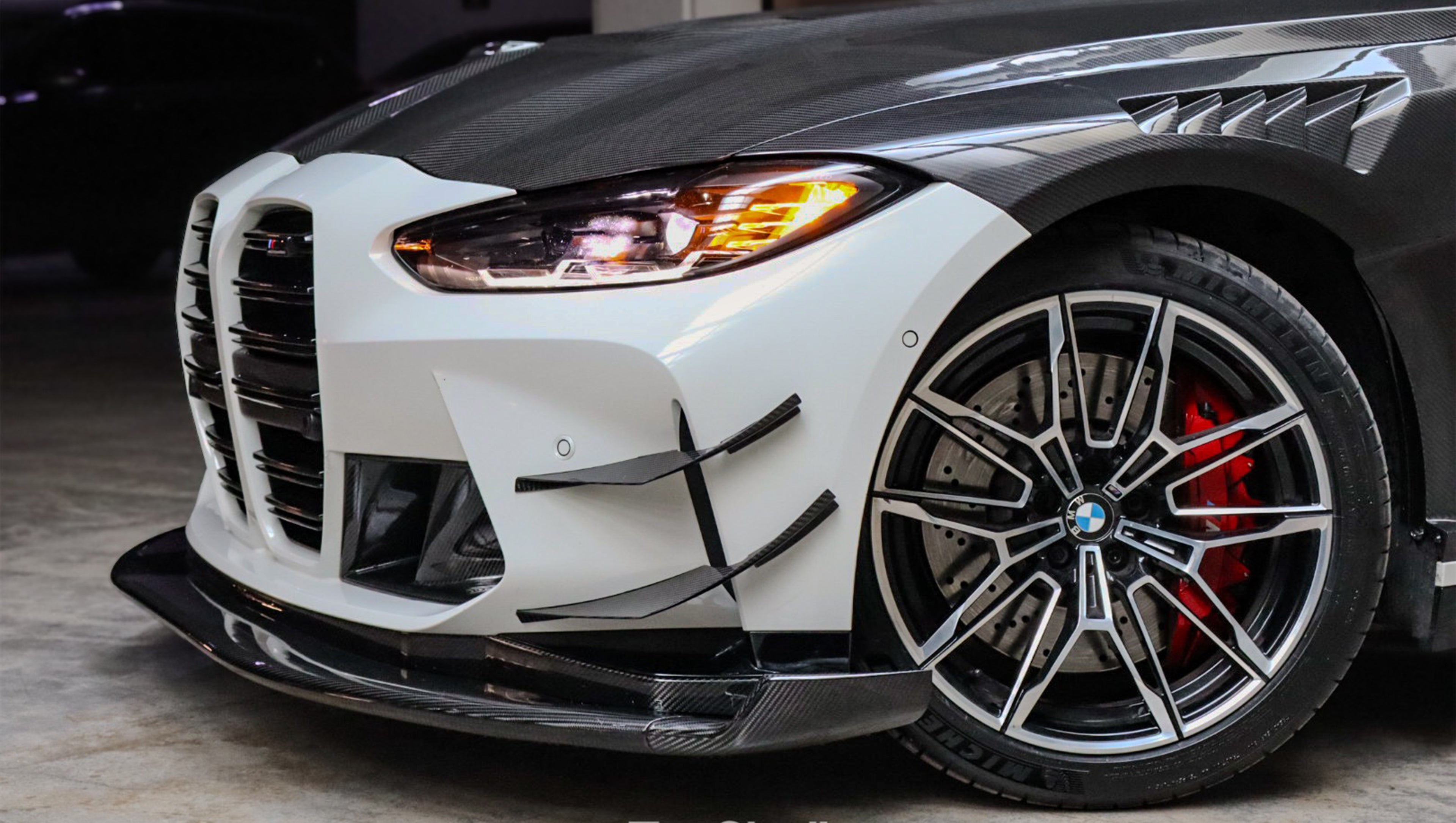 White BMW G82 M4 featuring a forged carbon fiber hood, front splitter, side canards, and aggressive aero styling parked inside a modern underground garage.