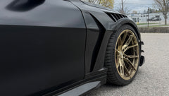 Close-up of a matte black BMW G82 M4 featuring REVOZPORT carbon fiber vented fenders, carbon side skirts, bronze forged wheels, and red performance brakes.