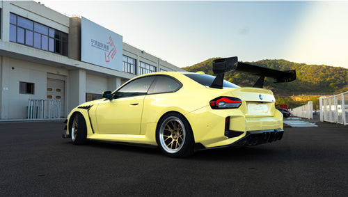 Pastel yellow BMW G82 M4 with carbon fiber front splitter, hood vents, side canards, bronze wheels, and large rear wing parked at a race track facility during golden hour.