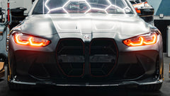 Matte black BMW G82 M4 with carbon fiber front splitter, side skirts, vented hood, bronze wheels, and oversized rear wing parked on a city street at night with headlights on.
