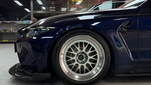 Dark blue BMW G82 M4 equipped with carbon fiber front splitter, side skirts, vented fenders, and a large rear wing, parked inside a modern luxury garage.