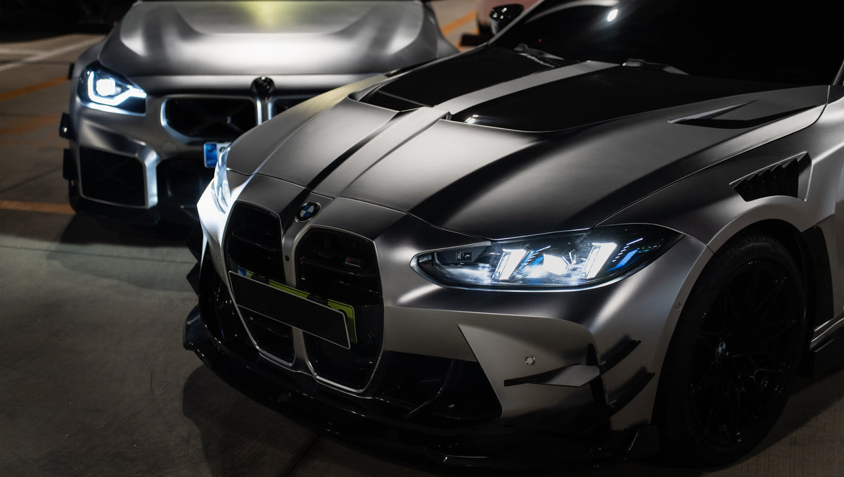 Matte gray BMW G82 M4 featuring an aggressive REVOZPORT carbon fiber aero kit with vented hood, front canards, wide fender vents, and illuminated LED headlights in a nighttime parking garage.