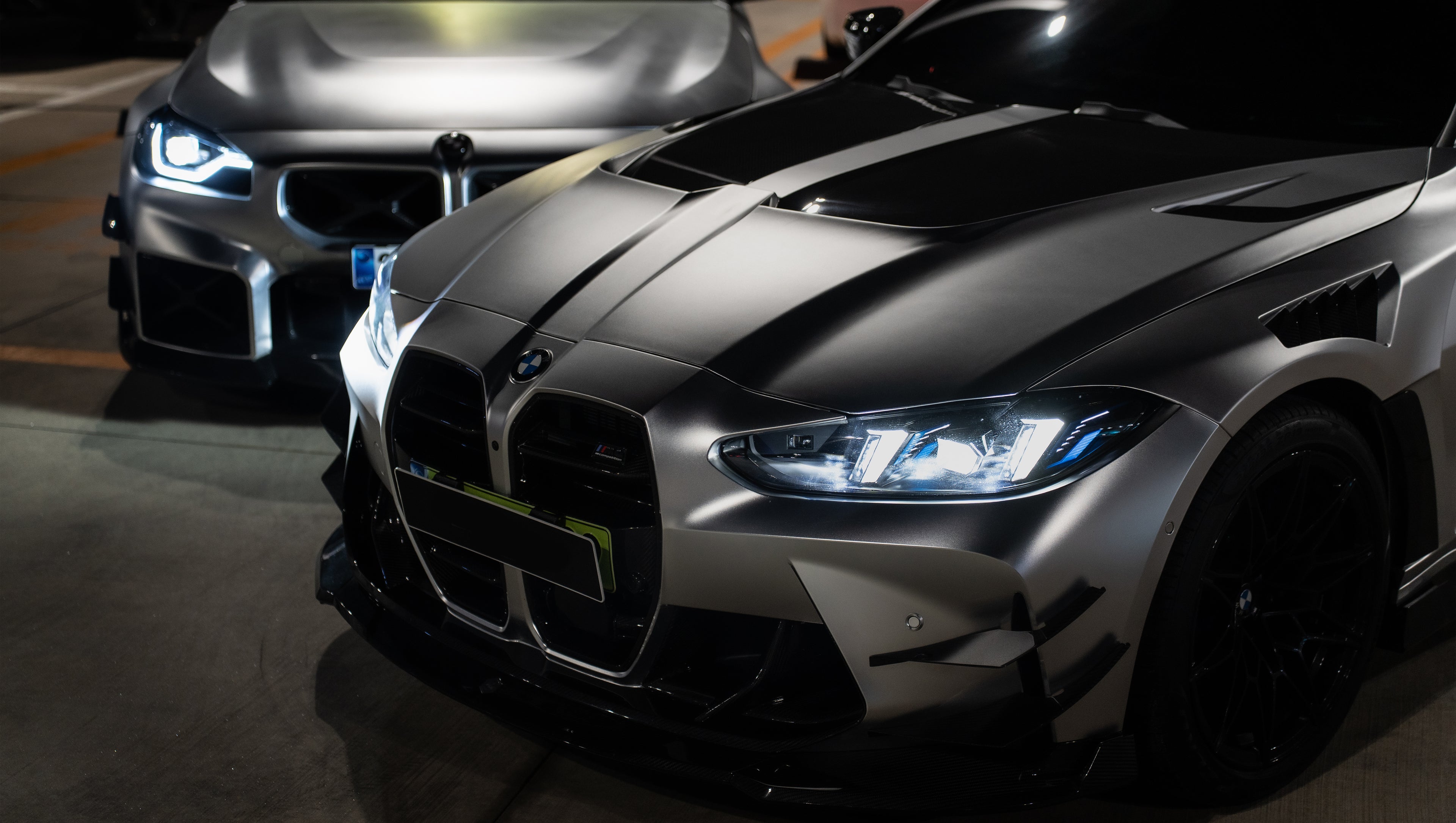 Matte gray BMW G82 M4 featuring an aggressive REVOZPORT carbon fiber aero kit with vented hood, front canards, wide fender vents, and illuminated LED headlights in a nighttime parking garage.