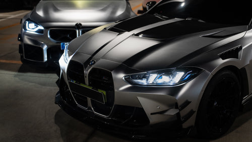 Matte gray BMW G82 M4 featuring an aggressive REVOZPORT carbon fiber aero kit with vented hood, front canards, wide fender vents, and illuminated LED headlights in a nighttime parking garage.