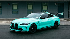 Mint green BMW G82 M4 with carbon fiber front splitter, side skirts, and large rear wing parked on a wet urban street outside a modern building.