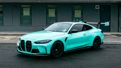 Mint green BMW G82 M4 with carbon fiber front splitter, side skirts, and large rear wing parked on a wet urban street outside a modern building.