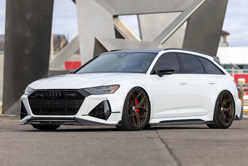 White Audi RS6 Avant featuring a REVOZPORT carbon fiber body kit with front lip, side skirts, rear diffuser, and lowered stance parked in a modern urban waterfront setting.