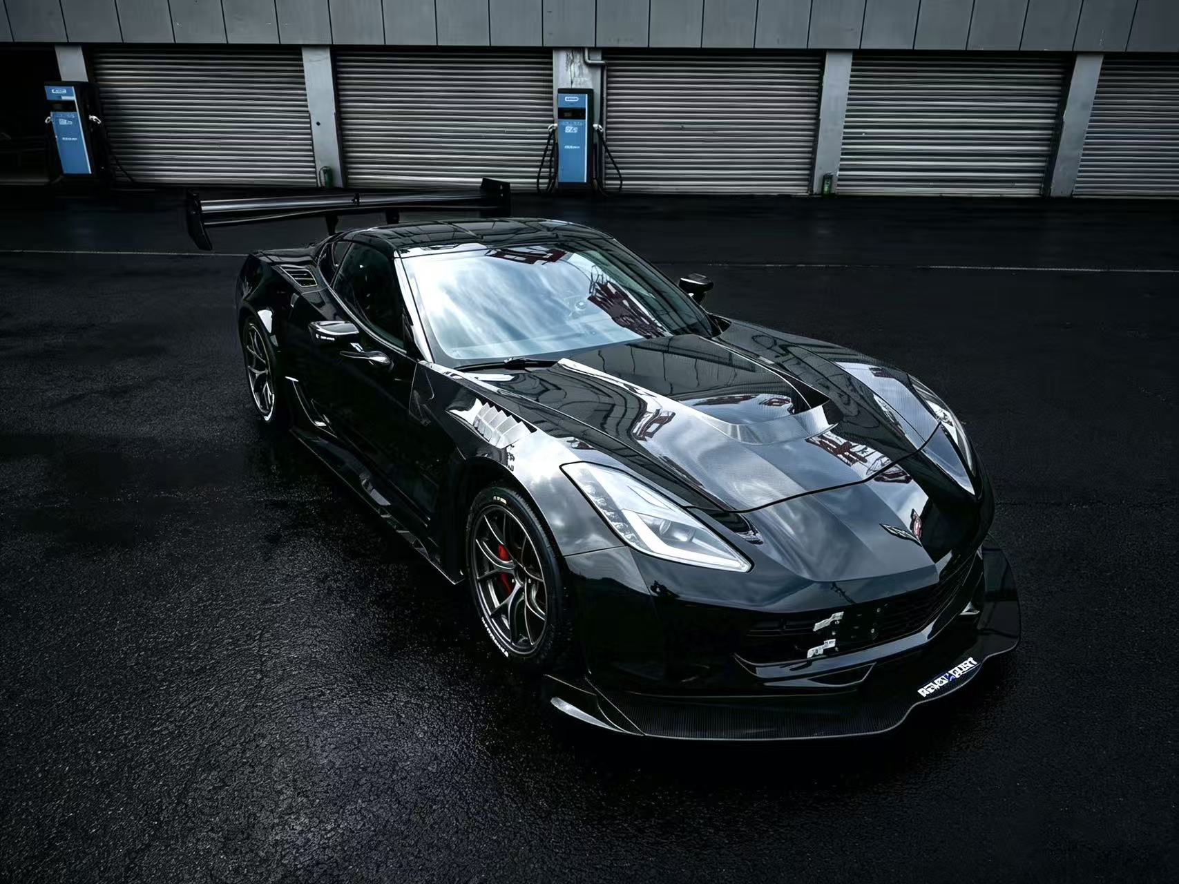 Black Chevrolet Corvette race car parked on a wet industrial track surface, featuring an aggressive wide-body kit, large rear wing, carbon fiber aerodynamic components, and lightweight performance wheels with red brake calipers. The moody urban backdrop and reflective pavement emphasize the car’s high-performance motorsport styling.