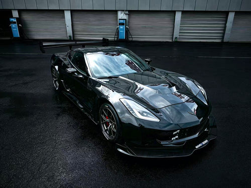 Black Chevrolet Corvette race car parked on a wet industrial track surface, featuring an aggressive wide-body kit, large rear wing, carbon fiber aerodynamic components, and lightweight performance wheels with red brake calipers. The moody urban backdrop and reflective pavement emphasize the car’s high-performance motorsport styling.