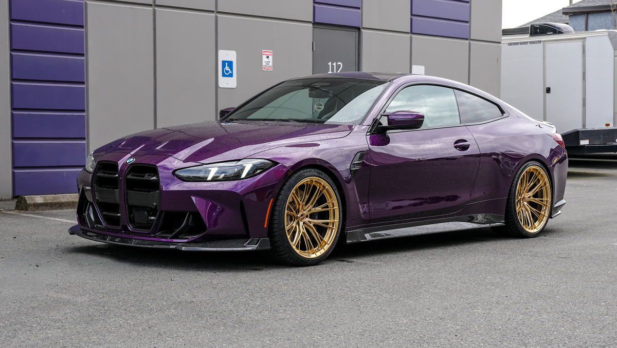Purple BMW G82 M4 featuring a REVOZPORT carbon fiber body kit with front lip, side skirts, vented fenders, and gold wheels parked outside a modern industrial building.
