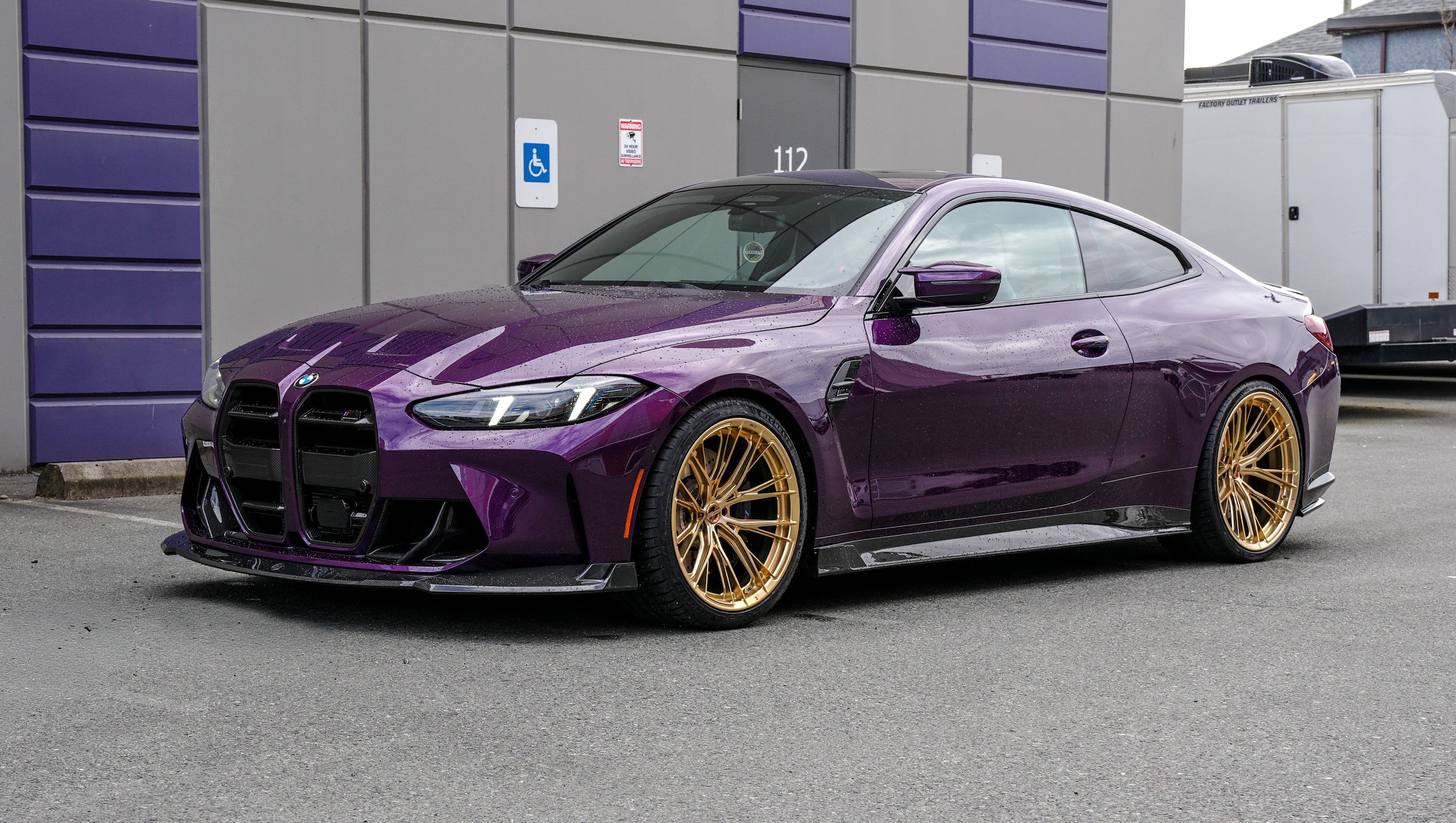 Purple BMW G82 M4 featuring a REVOZPORT carbon fiber body kit with front lip, side skirts, vented fenders, and gold wheels parked outside a modern industrial building.