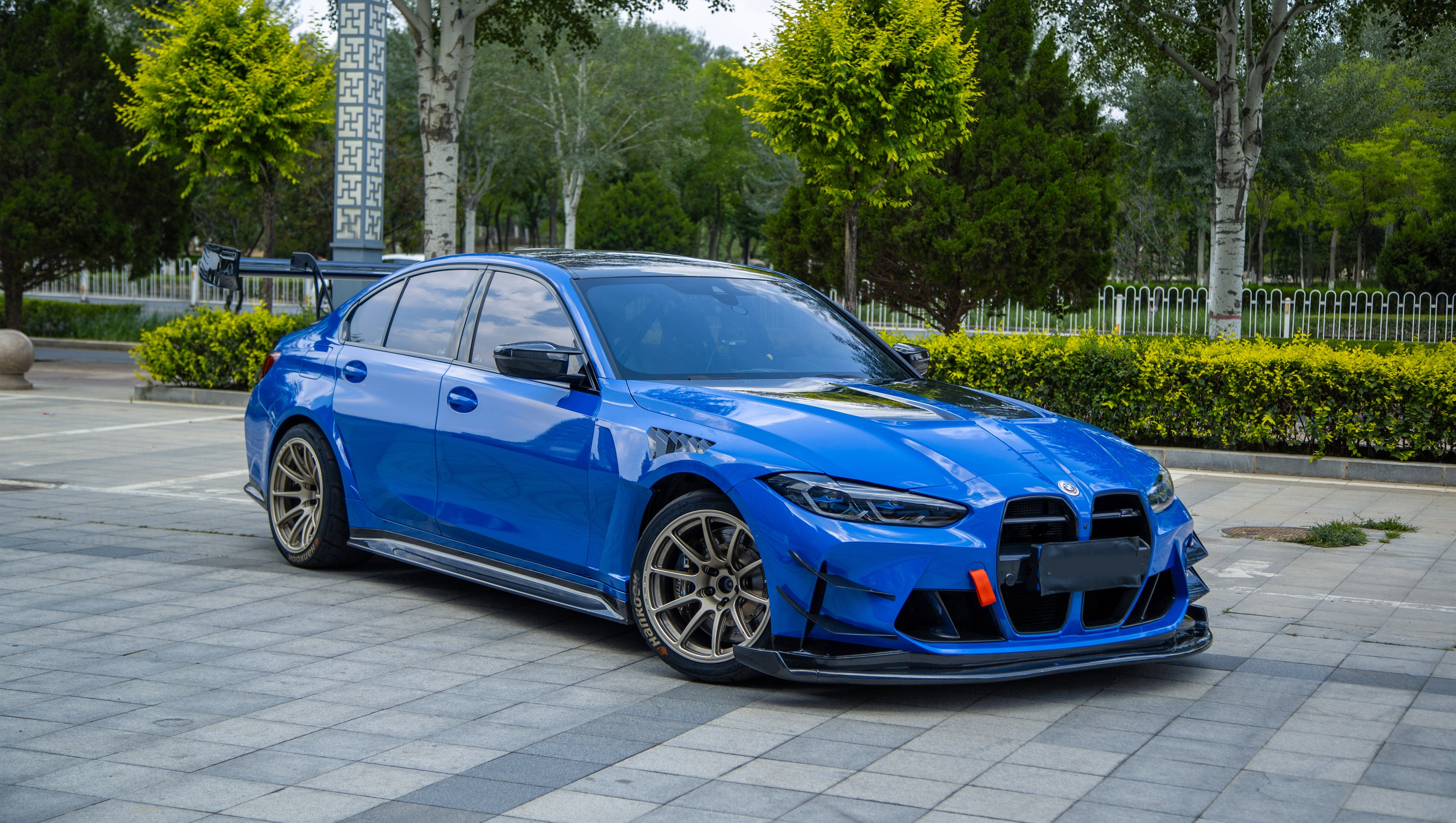 Blue BMW G80 M3 with REVOZPORT carbon fiber front splitter, side skirts, vented fenders, and large rear wing parked in a landscaped outdoor plaza.