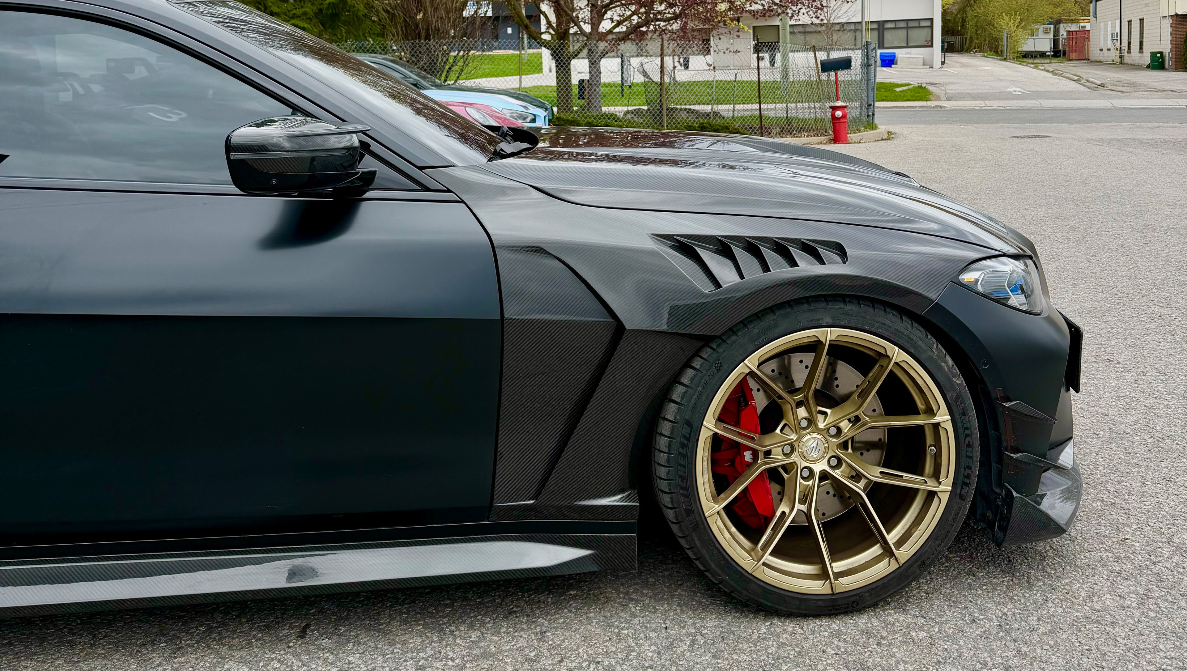 Close-up of a matte black BMW G82 M4 featuring REVOZPORT carbon fiber vented fenders, carbon side skirts, bronze forged wheels, and red performance brakes.
