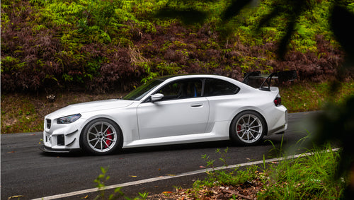 White widebody BMW G82 M4 with aggressive front bumper, canards, deep front splitter, large rear wing, and yellow LED headlights parked on a wet road surrounded by dense tropical forest.