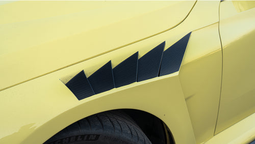 Pastel yellow BMW G82 M4 with carbon fiber front splitter, hood vents, side canards, bronze wheels, and large rear wing parked at a race track facility during golden hour.