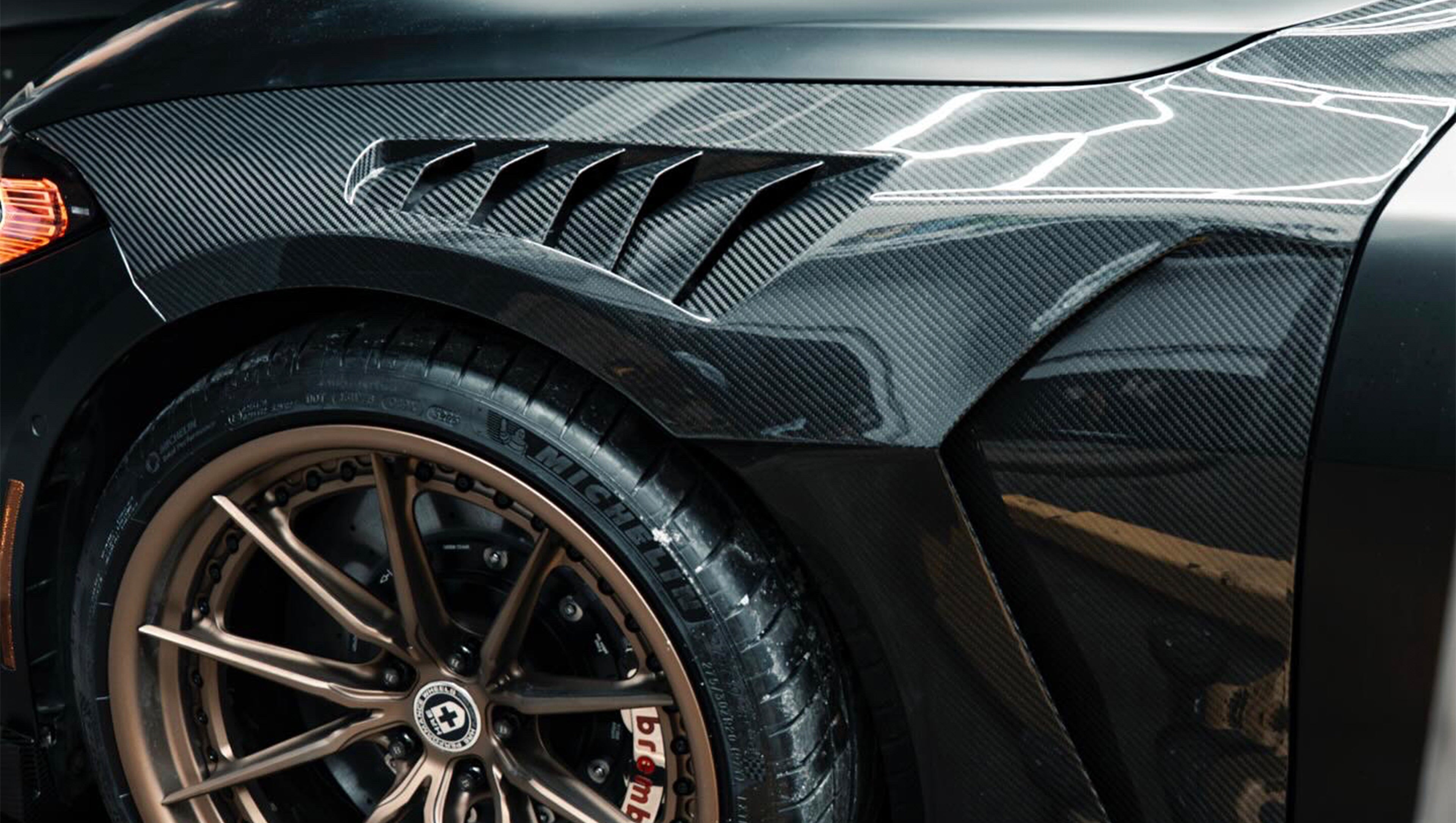 Matte black BMW G82 M4 with carbon fiber front splitter, side skirts, vented hood, bronze wheels, and oversized rear wing parked on a city street at night with headlights on.