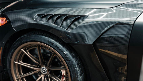 Matte black BMW G82 M4 with carbon fiber front splitter, side skirts, vented hood, bronze wheels, and oversized rear wing parked on a city street at night with headlights on.
