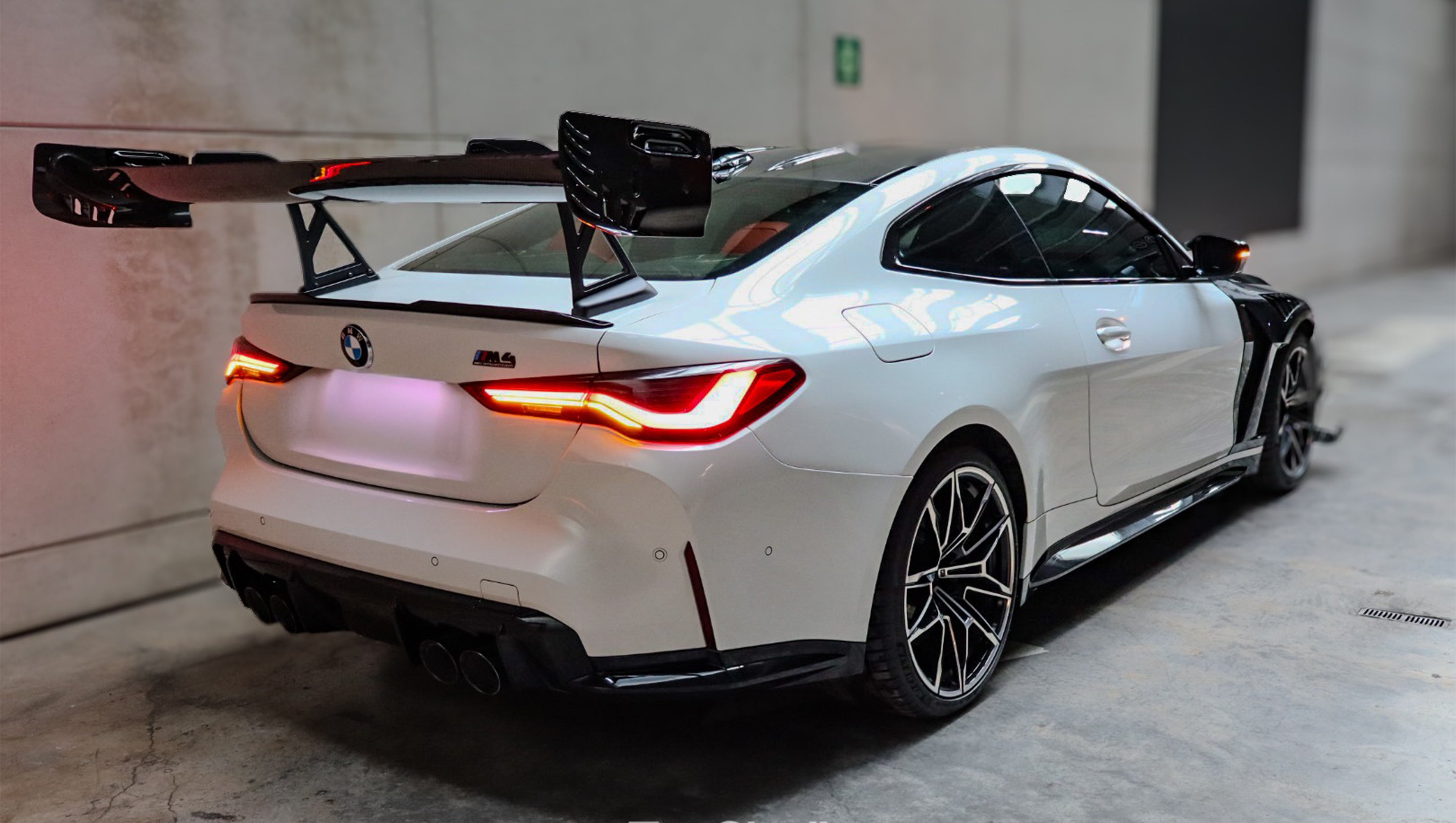 White BMW G82 M4 featuring a forged carbon fiber hood, front splitter, side canards, and aggressive aero styling parked inside a modern underground garage.