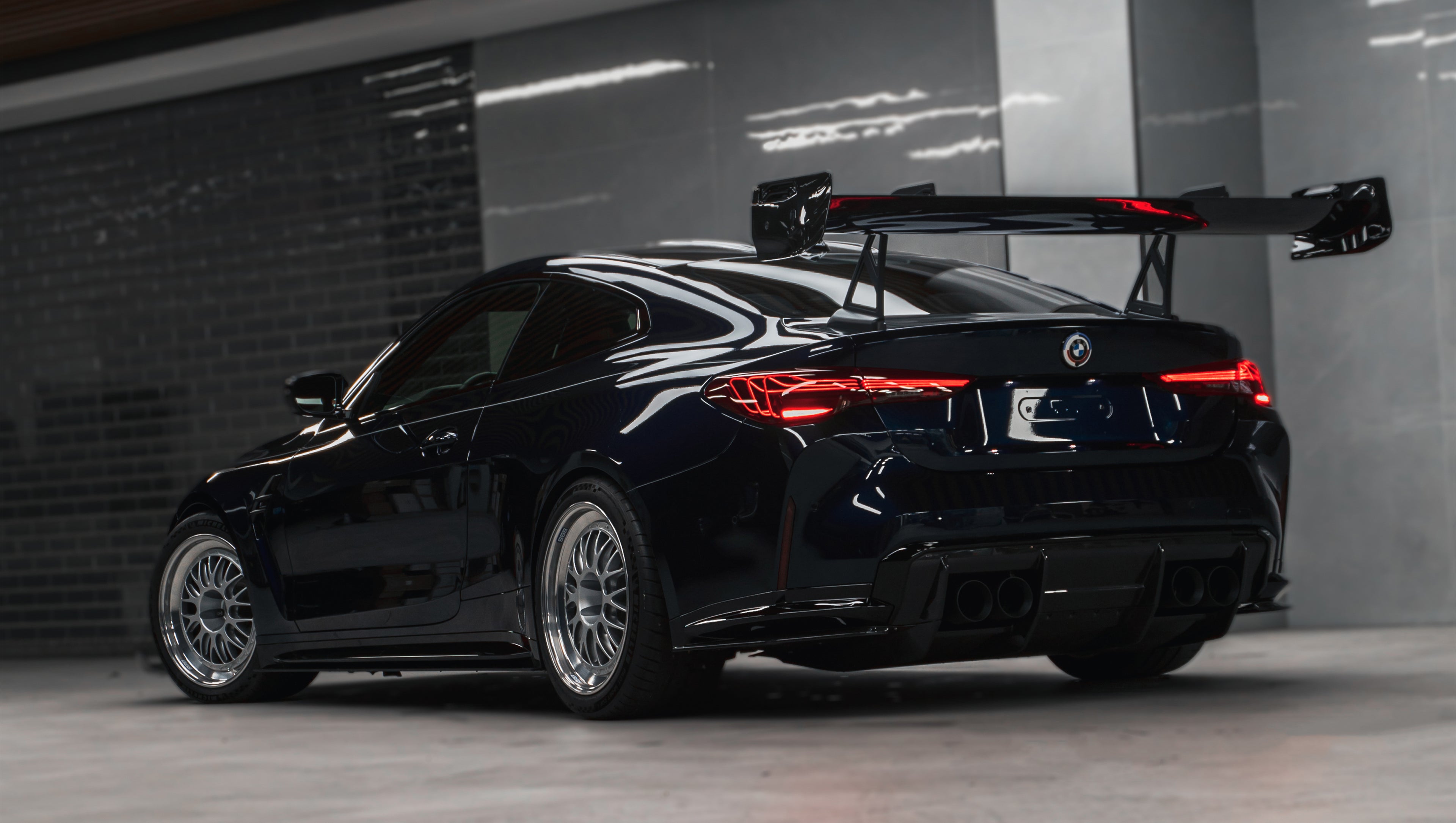 Dark blue BMW G82 M4 equipped with carbon fiber front splitter, side skirts, vented fenders, and a large rear wing, parked inside a modern luxury garage.