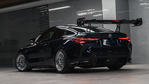 Dark blue BMW G82 M4 equipped with carbon fiber front splitter, side skirts, vented fenders, and a large rear wing, parked inside a modern luxury garage.