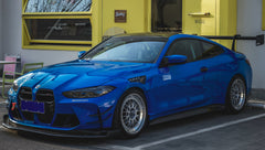 Blue BMW G82 M4 with carbon fiber front splitter, side skirts, rear wing, and aftermarket wheels parked in a modern industrial courtyard with colorful architectural walls.