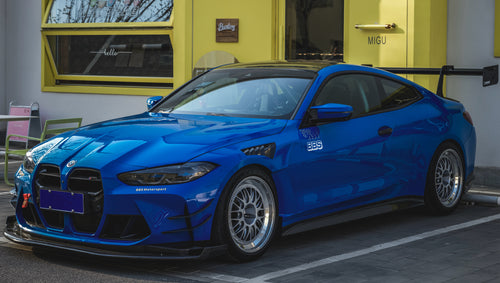 Blue BMW G82 M4 with carbon fiber front splitter, side skirts, rear wing, and aftermarket wheels parked in a modern industrial courtyard with colorful architectural walls.