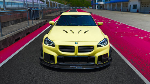 Yellow BMW G82 M4 with aggressive carbon fiber front splitter, vented hood, side canards, and bronze wheels parked on a race track pit lane.