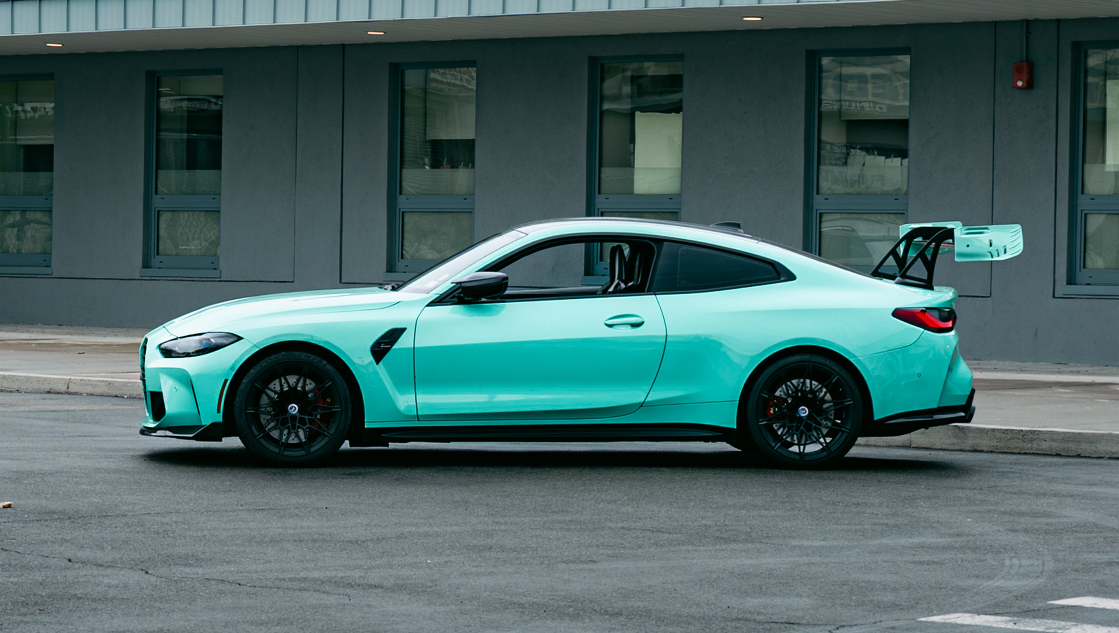 Mint green BMW G82 M4 with carbon fiber front splitter, side skirts, and large rear wing parked on a wet urban street outside a modern building.