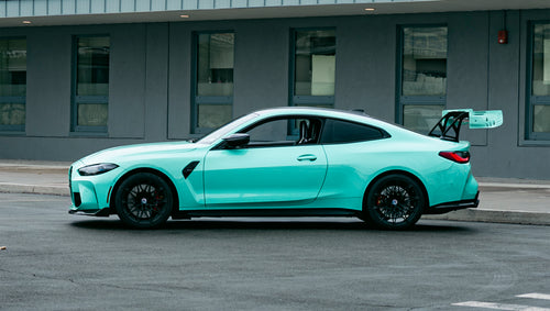 Mint green BMW G82 M4 with carbon fiber front splitter, side skirts, and large rear wing parked on a wet urban street outside a modern building.